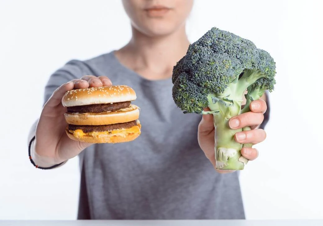 Žena u jednoj ruci drži brokoli a u drugoj hamburger.